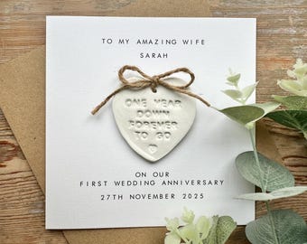 May include: White card with a white heart-shaped ornament attached with twine. The ornament has the text "One Year Down Forever To Go" on it. The card has the text "To My Amazing Wife Sarah On Our First Wedding Anniversary 27th November 2025".