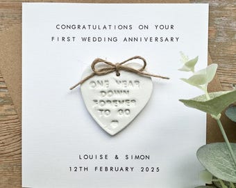 May include: White card with the text "Congratulations on your first wedding anniversary." A white heart-shaped ornament with the text "One year down forever to go" is attached to the card. The card also includes the names "Louise & Simon" and the date "12th February 2025".