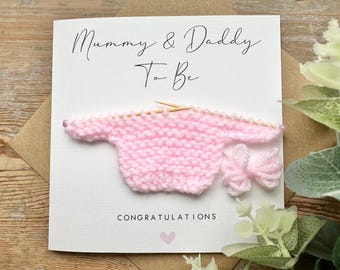 May include: A pink knitted baby sweater and matching hat with the text "Mummy & Daddy To Be Congratulations" on a white card.