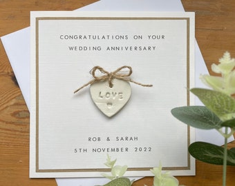 May include: A wedding anniversary card with the text "CONGRATULATIONS ON YOUR WEDDING ANNIVERSARY". It features a heart-shaped ornament with the word "LOVE" and a twine bow. The names "ROB & SARAH" and the date "5TH NOVEMBER 2022" are also printed.