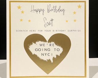 May include: A white card with gold trim and stars. The card says "Happy Birthday Scott" and "Scratch here for your birthday surprise". A gold heart with a white scratch-off area says "We're going to NYC!"