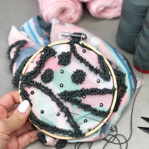 以下が含まれることがあります： ピンク、グリーン、ブラックのパターンが施された円形の刺繍枠のクローズアップ。生地は、ブラックの糸のポンポンとブラックのラインストーンで飾られています。枠の中に針と糸が見えます。