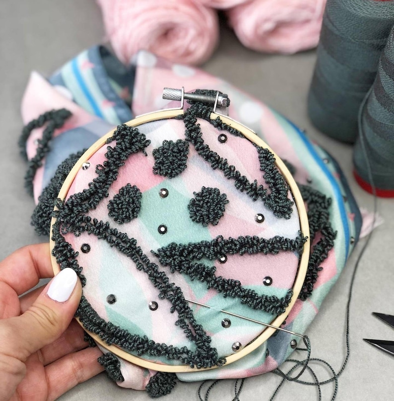 以下が含まれることがあります： ピンク、グリーン、ブラックのパターンが施された円形の刺繍枠のクローズアップ。生地は、ブラックの糸のポンポンとブラックのラインストーンで飾られています。枠の中に針と糸が見えます。