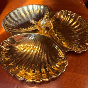 May include: A gold-colored metal serving dish shaped like three conjoined scallop shells. The dish features a central decorative element in the form of a small squirrel figure. The surface has a reflective, polished finish with scalloped edges.