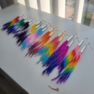 May include: A collection of beaded fringe earrings in various colors, including rainbow, pink, blue, and yellow. Each earring features a silver or gold-toned metal post and a cascade of colorful beads, creating a vibrant and eye-catching accessory.