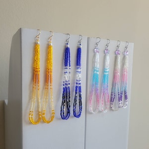 May include: Beaded dangle earrings in shades of yellow, blue, and a pink-purple gradient. The earrings are displayed on a white surface. The intricate beadwork and delicate design of the earrings are clearly visible.