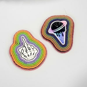 May include: Two beaded patches with rainbow-colored borders. One patch features a white skeleton hand making a middle finger gesture. The other patch displays a black ice cream cone graphic with a planet ring design against a starry background.