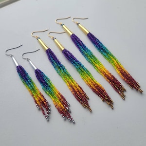 May include: Six pairs of beaded dangle earrings. The earrings feature a rainbow gradient of colors, transitioning from purple to red. The top portion of the earrings is gold or silver, with a hook for the ear. The beads are small and create a fringed effect.