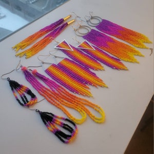 May include: A collection of beaded earrings in various styles and colors. The earrings feature long fringe designs in shades of purple, pink, orange, and yellow. Some earrings have silver or gold-colored metal accents, while others are black.