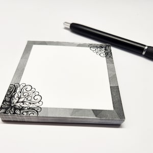 May include: A gray and white notepad with a decorative black border. The notepad has a blank white space in the center for writing. A black pen is lying on the white surface next to the notepad.