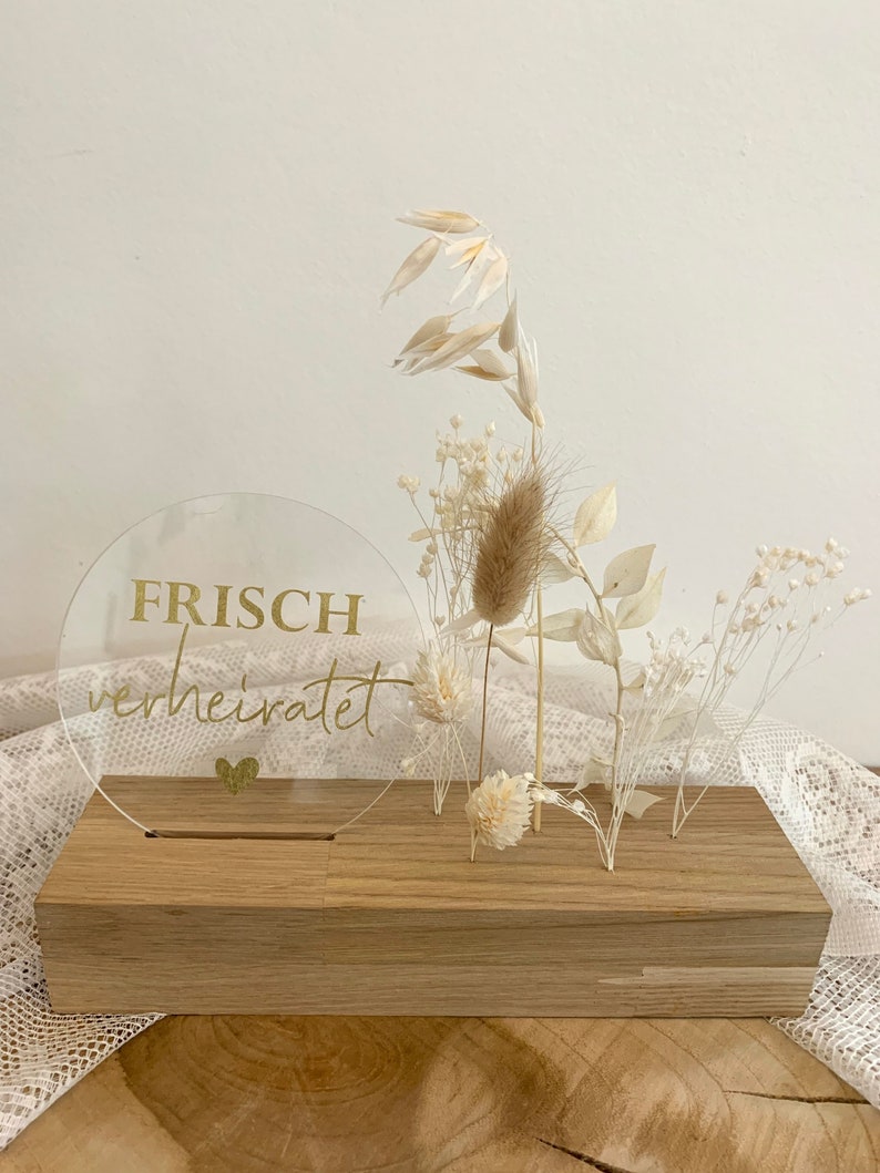 Może przedstawiać: Przezroczysty okrągły akryl z napisem "FRISCH verheiratet" złotymi literami, stojący na drewnianej podstawie z suszonymi kwiatami i trawami.