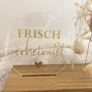 Może przedstawiać: Przezroczysty znak akrylowy z napisem "FRISCH verheiratet" złotymi literami i złotym sercem. Znak stoi na drewnianej podstawie.