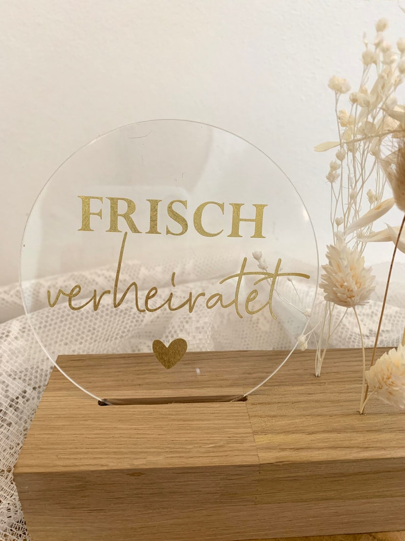 Może przedstawiać: Przezroczysty znak akrylowy z napisem "FRISCH verheiratet" złotymi literami i złotym sercem. Znak stoi na drewnianej podstawie.