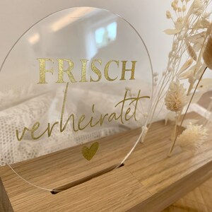Może przedstawiać: Okrągły znak z przezroczystego akrylu z złotym napisem "FRISCH verheiratet" i małym złotym sercem pod tekstem. Znak stoi na drewnianej podstawie.