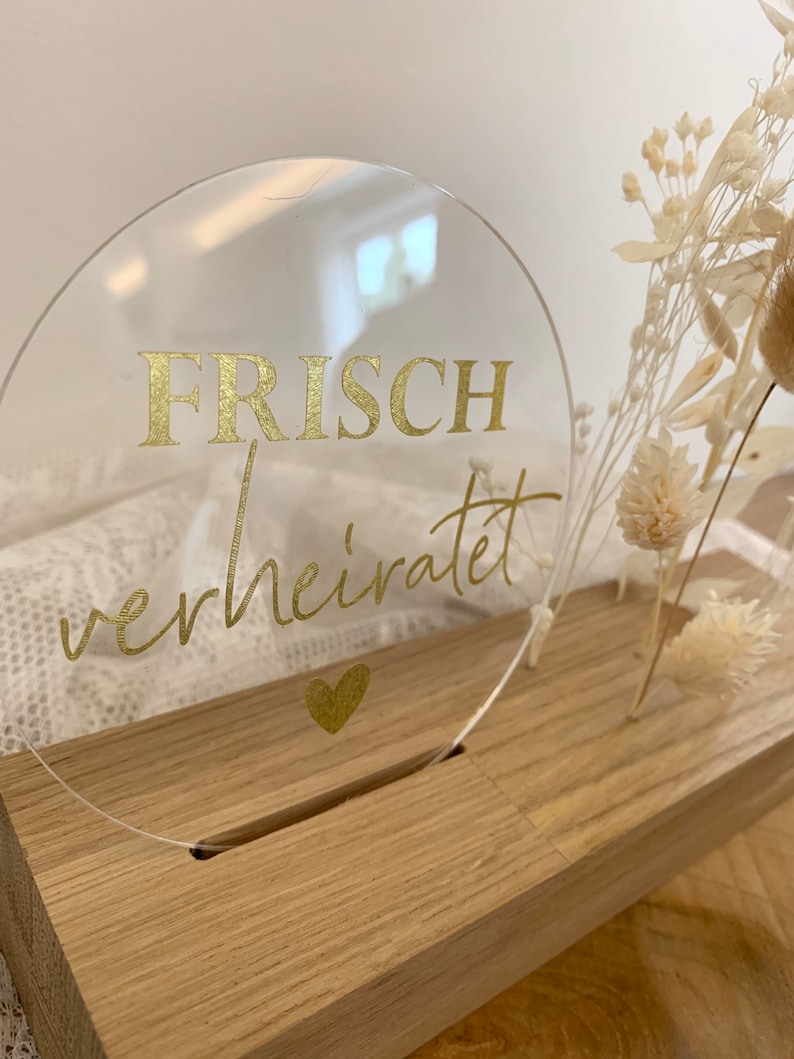 Może przedstawiać: Okrągły znak z przezroczystego akrylu z złotym napisem "FRISCH verheiratet" i małym złotym sercem pod tekstem. Znak stoi na drewnianej podstawie.