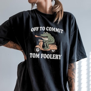 May include: Black t-shirt with the text "OFF TO COMMIT TOM FOOLERY." A cartoon frog with a long tongue is riding a peach-colored scooter. The frog is green with black spots. The text is in a white, bold font.