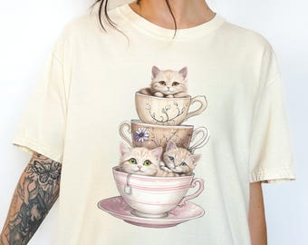 May include: Cream-colored t-shirt featuring a whimsical design of three kittens stacked in teacups. The teacups are decorated with floral patterns and a teabag. The kittens have light brown fur and different colored eyes. The design is centered on the front of the shirt.