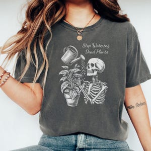 May include: Dark gray t-shirt featuring a white graphic of a skeleton watering a potted plant with a watering can. The text "Stop Watering Dead Plants" is above the skeleton. The shirt is a casual, unisex style.