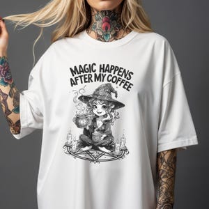 Op de afbeelding: Wit T-shirt met een zwart-wit illustratie van een heks die een koffiekopje vasthoudt, omringd door kaarsen. De tekst "MAGIC HAPPENS AFTER MY COFFEE" is boven de illustratie gedrukt. Het shirt heeft korte mouwen.