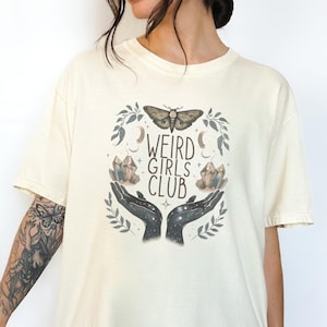 Op de afbeelding: Crèmekleurig T-shirt met een grafisch ontwerp. Het ontwerp bevat de tekst "WEIRD GIRLS CLUB", een mot, kristallen, handen, manen en sterren, omringd door een bladerkrans. Het shirt heeft korte mouwen.