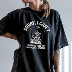 以下が含まれることがあります： 白いイラストが描かれた黒いTシャツ。ガチョウがスクーターに乗っており、上部に「SORRY, I CAN'T」、下部に「I HAVE TO FEED MY SOURDOUGH STARTER」と書かれています。Tシャツはチャコールグレーです。