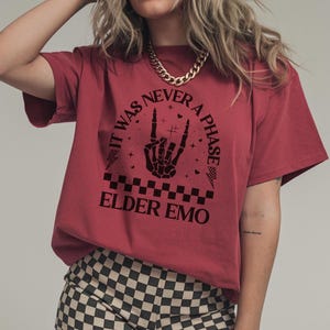 May include: Red t-shirt with the text "IT WAS NEVER A PHASE" in a circular design, featuring a skeleton hand making a rock and roll gesture. The shirt also has the words "ELDER EMO" below the design.