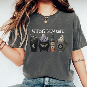 以下が含まれることがあります： 「Witch's Brew Café」の文字と、ハロウィーンをテーマにした4つのイラストがプリントされた濃いグレーのTシャツ。イラストには、飲み物、大釜、ポーションボトル、お化けのマグカップが含まれています。
