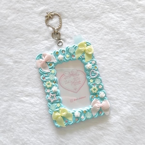 May include: A rectangular light blue card holder featuring a textured whipped cream-like border decorated with small pink and yellow bows, white floral accents, pearl-like beads, and star shapes. A silver metal keychain is attached to the top.