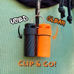May include: A black and an orange cylindrical container with a spiral design, attached to a keyring. The words "USED", "CLEAN", and "CLIP & GO!" are written on the image.