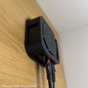 Op de afbeelding: Een zwarte, aan de muur gemonteerde apparaathouder met een gebogen ontwerp, bevestigd aan een houten oppervlak. De houder heeft een cirkelvormige bovenkant met een logo en sleuven. Kabels zijn aangesloten op het apparaat. De tekst "DEVICE NOT INCLUDED!" staat onderaan.