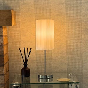 May include: A chrome table lamp with a white pleated cylindrical lampshade. The lamp is on a glass table with a brown glass bottle and diffuser sticks.