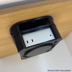 May include: Black, 3D-printed device holder, mounted under a wooden surface. The holder encases a silver device with two small, vertical slots and a circular, vented base. The text "DEVICE NOT INCLUDED!" is at the bottom.
