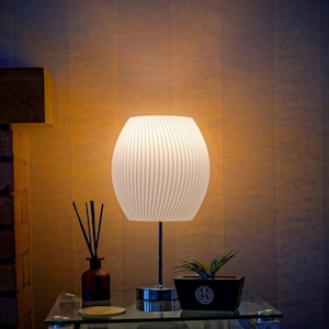 May include: A white ribbed lampshade with a silver base sits on a glass table with a small potted plant and a brown diffuser bottle with sticks.