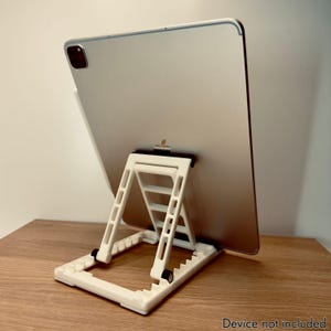 May include: A white tablet stand with a triangular design, supporting a silver tablet. The stand has a foldable design and is made of plastic. The text "Device not included" is visible at the bottom.