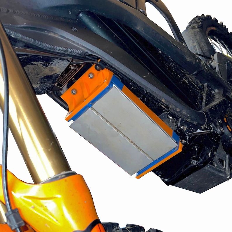 May include: Detail of an electric mountain bike's battery pack. The silver battery is framed by orange and blue components, attached to the black frame. The front suspension fork is gold. The bike is designed for off-road use.