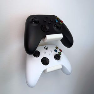 May include: A white wall-mounted dual controller holder featuring two stacked slots. The holder supports one black and one white gaming controller. The text "Controllers not included!" is printed in the bottom right corner of the image.