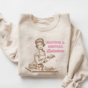 Peut inclure: Un sweat-shirt beige avec une illustration r&eacute;tro d'une femme en train de faire des tartes. Le texte "HAVING A MENTAL BAKEDOWN" est imprim&eacute; en lettres brunes et roses.