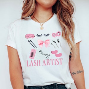 Pu&ograve; includere: Maglietta bianca con un fiocco rosa e una grafica di strumenti per l'artista delle ciglia. Il testo "LASH ARTIST" &egrave; stampato in lettere rosa sulla maglietta.