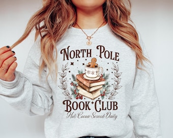 May include: A gray sweatshirt with the text "North Pole Book Club" and "Hot Cocoa Served Daily". A gingerbread man is sitting on a stack of books with a mug of hot cocoa.