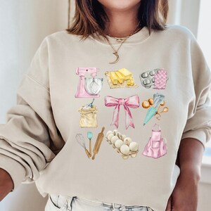 May include: A light beige sweatshirt with a watercolor design of baking supplies. The design includes a pink stand mixer, a rolling pin, a whisk, a bag of flour, a pink apron, a bowl of eggs, a bow, a cupcake liner, a piping bag, and a stick of butter.