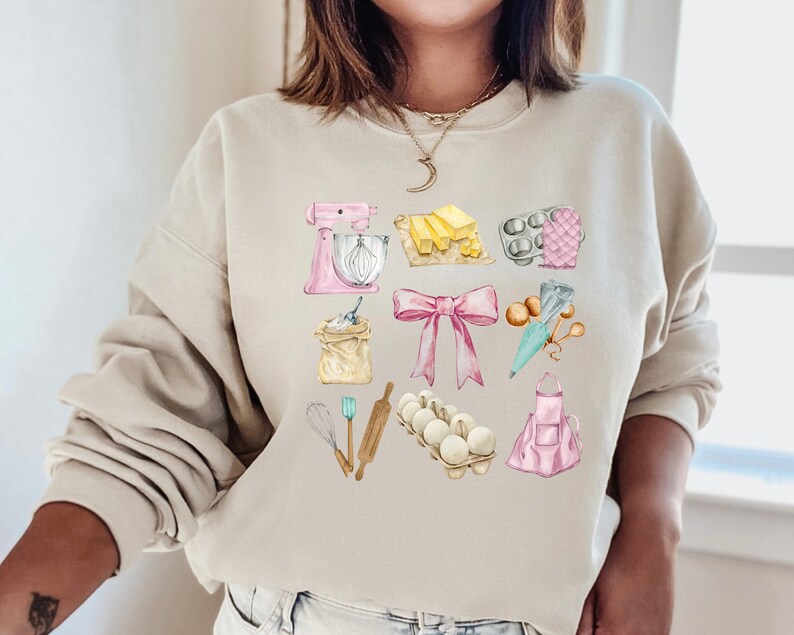 May include: A light beige sweatshirt with a watercolor design of baking supplies. The design includes a pink stand mixer, a rolling pin, a whisk, a bag of flour, a pink apron, a bowl of eggs, a bow, a cupcake liner, a piping bag, and a stick of butter.