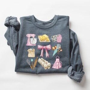 May include: A dark gray sweatshirt with a watercolor design of baking supplies. The design includes a pink stand mixer, a bag of flour, a pink bow, a carton of eggs, a rolling pin, a whisk, a pink apron, a piping bag, a muffin tin, and a stick of butter.