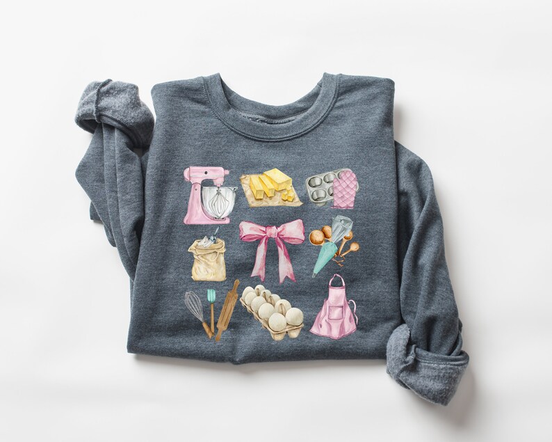 May include: A dark gray sweatshirt with a watercolor design of baking supplies. The design includes a pink stand mixer, a bag of flour, a pink bow, a carton of eggs, a rolling pin, a whisk, a pink apron, a piping bag, a muffin tin, and a stick of butter.