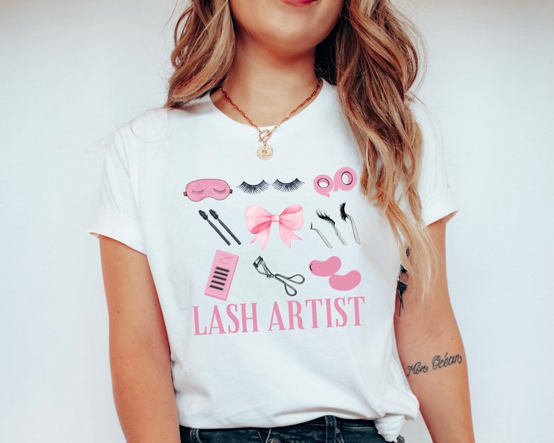Pu&ograve; includere: Maglietta bianca con un fiocco rosa e una grafica di strumenti per l'artista delle ciglia. Il testo "LASH ARTIST" &egrave; stampato in lettere rosa sulla maglietta.