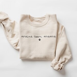 May include: A beige sweatshirt with the text "maine coon mama" and a paw print in black.