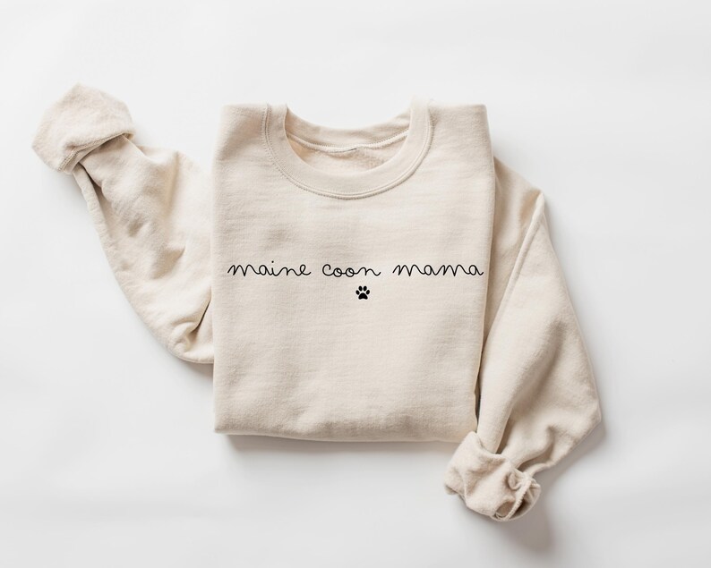 May include: A beige sweatshirt with the text "maine coon mama" and a paw print in black.