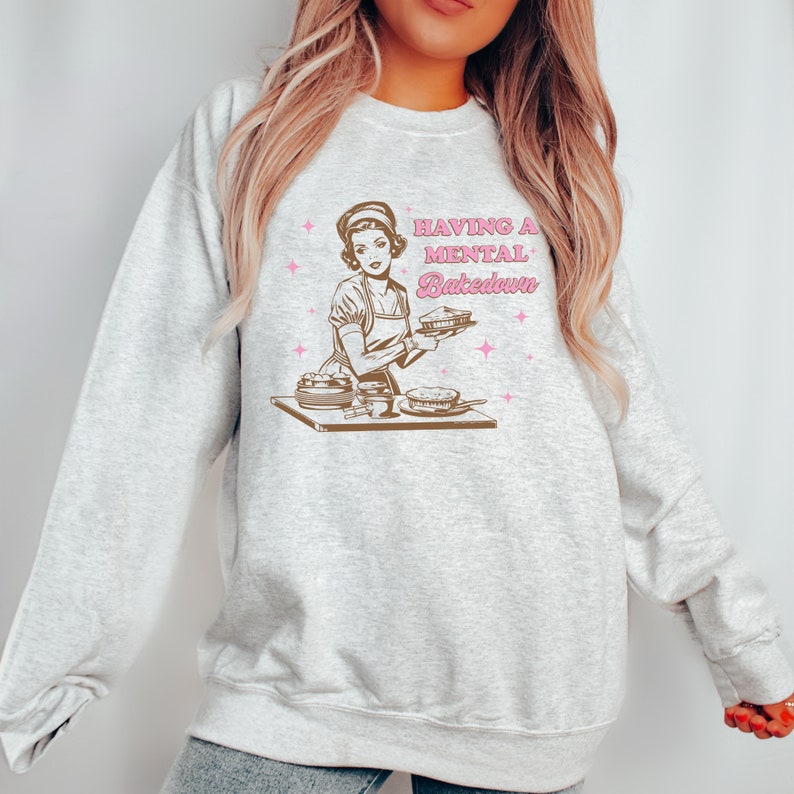 Peut inclure: Un sweat-shirt gris avec une illustration r&eacute;tro d'une femme en train de cuisiner. Le texte sur le sweat-shirt est "Having a Mental Bakedown".