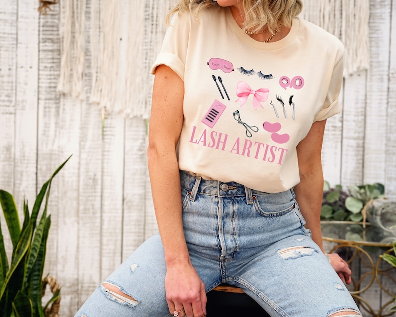 Pu&ograve; includere: Una maglietta beige chiaro con un design grafico di strumenti per l'estensione delle ciglia e il testo "Lash Artist".
