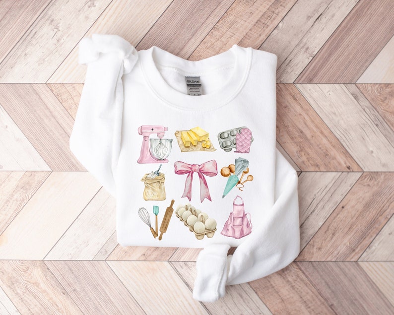 May include: White sweatshirt with a pink, blue, and yellow watercolor design of baking supplies, including a mixer, flour, butter, eggs, a rolling pin, a whisk, and an apron.