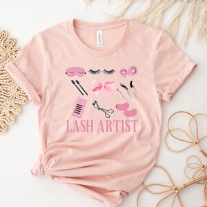 Pu&ograve; includere: Una maglietta rosa con un design grafico di strumenti per artisti di ciglia e il testo "LASH ARTIST".